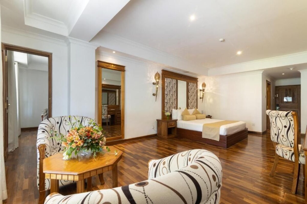 A hotel room with a bed, a dining table with chairs, a mirror, and a flower arrangement.