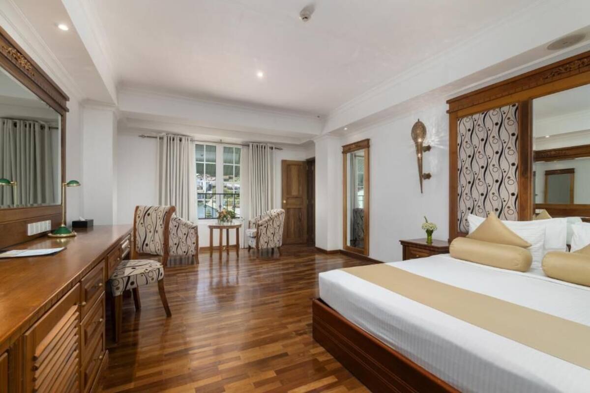 Ein Hotelzimmer mit einem großen Bett, einem Schreibtisch mit Lampe, zwei Stühlen, einem kleinen Tisch mit einer Vase und einem großen Spiegel.