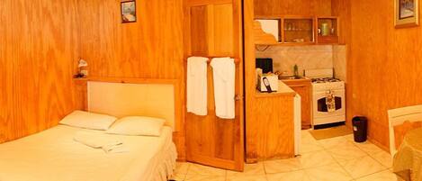 Una habitación compacta con una cama, una kitchenette pequeña y una puerta que da a otra habitación.