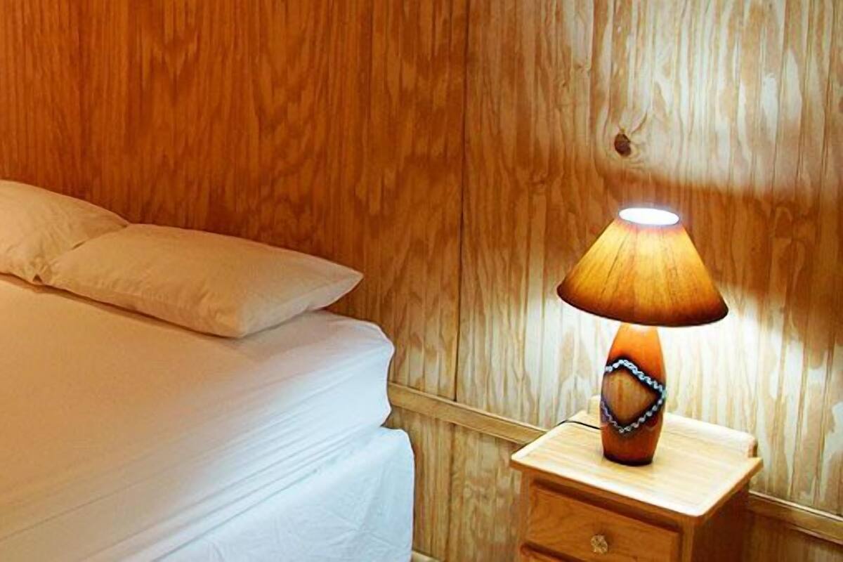 A neatly made bed with a towel in the shape of a swan, a bedside lamp, and wooden paneling on the walls.