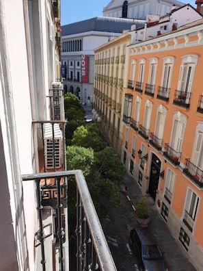 A narrow street with buildings on either side, featuring balconies and a small courtyard with a tree.