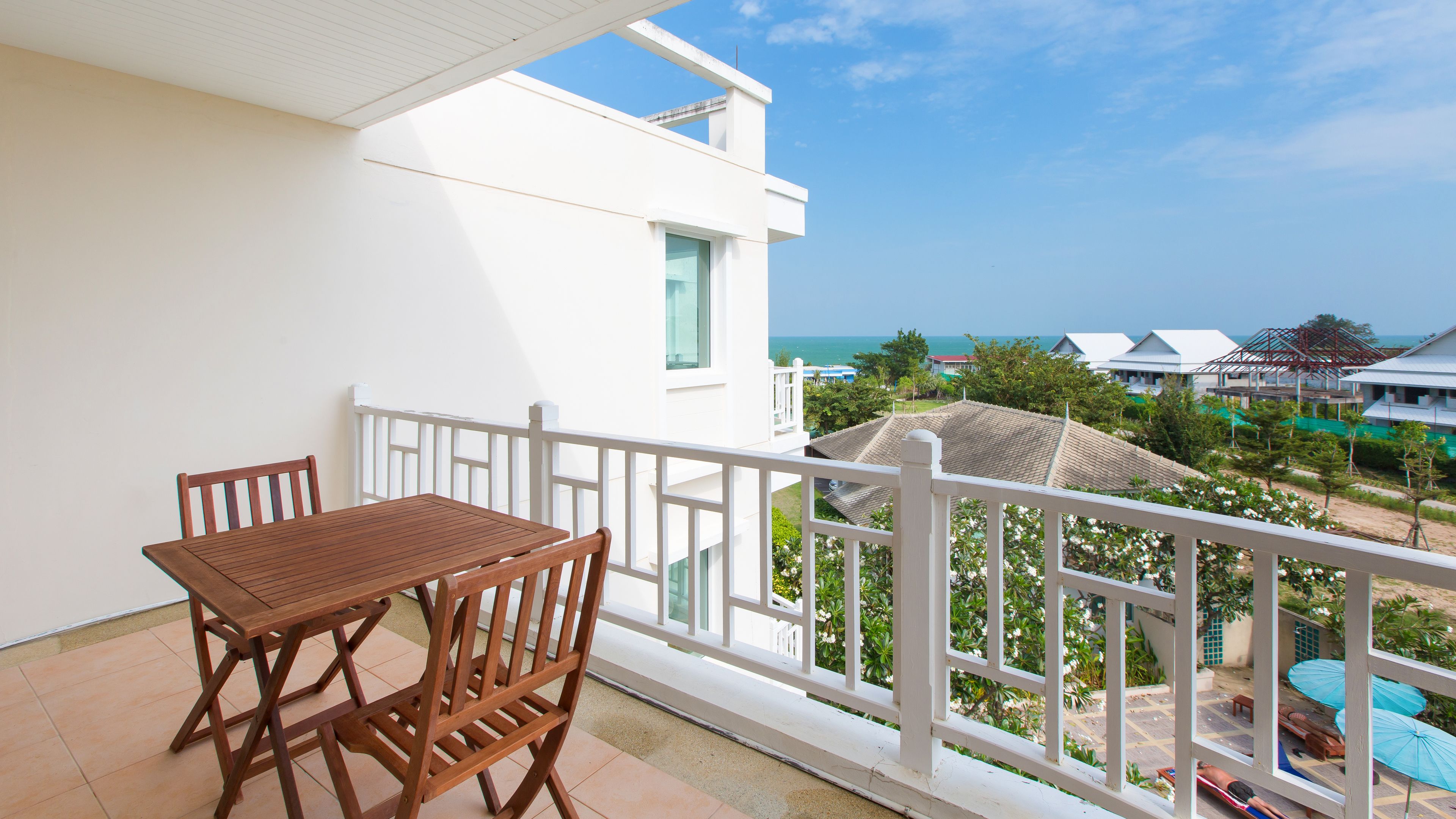 A balcony with a wooden table and chairs, overlooking a coastal area with buildings and a clear sky.