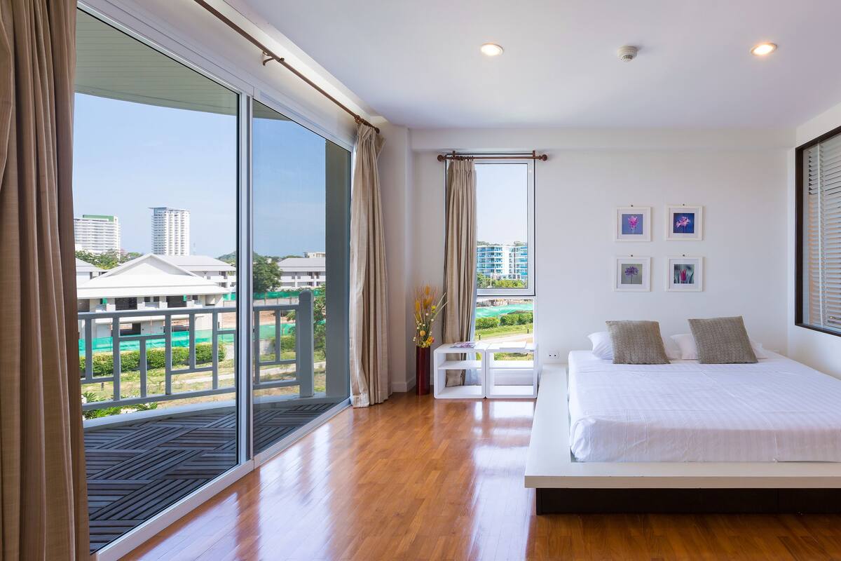 A modern bedroom with a large bed, a view of the city, and a balcony with a railing.