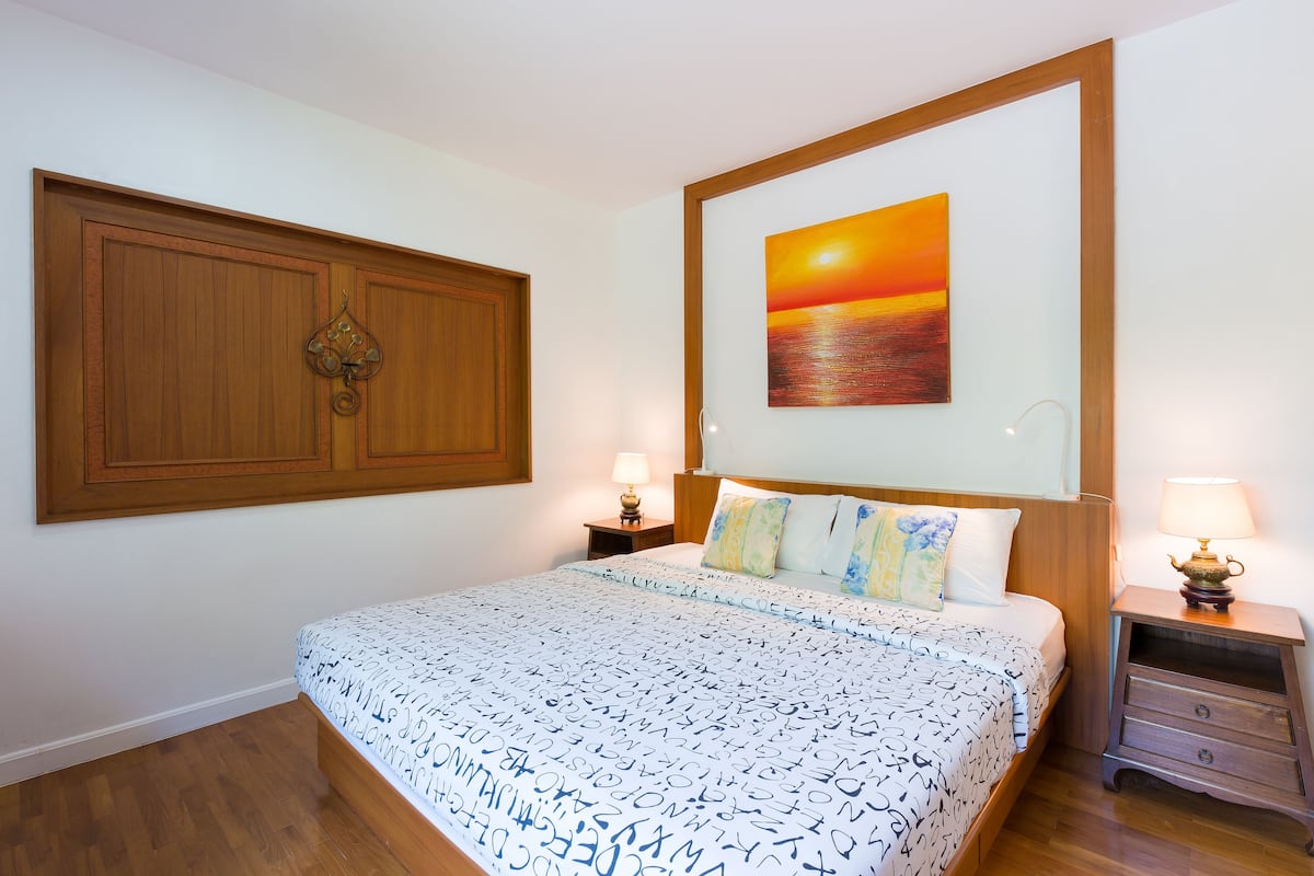 A neatly arranged bedroom with a large bed, a wooden wardrobe, a bedside table with a lamp, and a painting of a sunset on the wall.