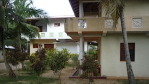 A building with a covered outdoor area, palm trees, and a sandy ground. - Beach Lagoon Guesthouse (Tangalle)
