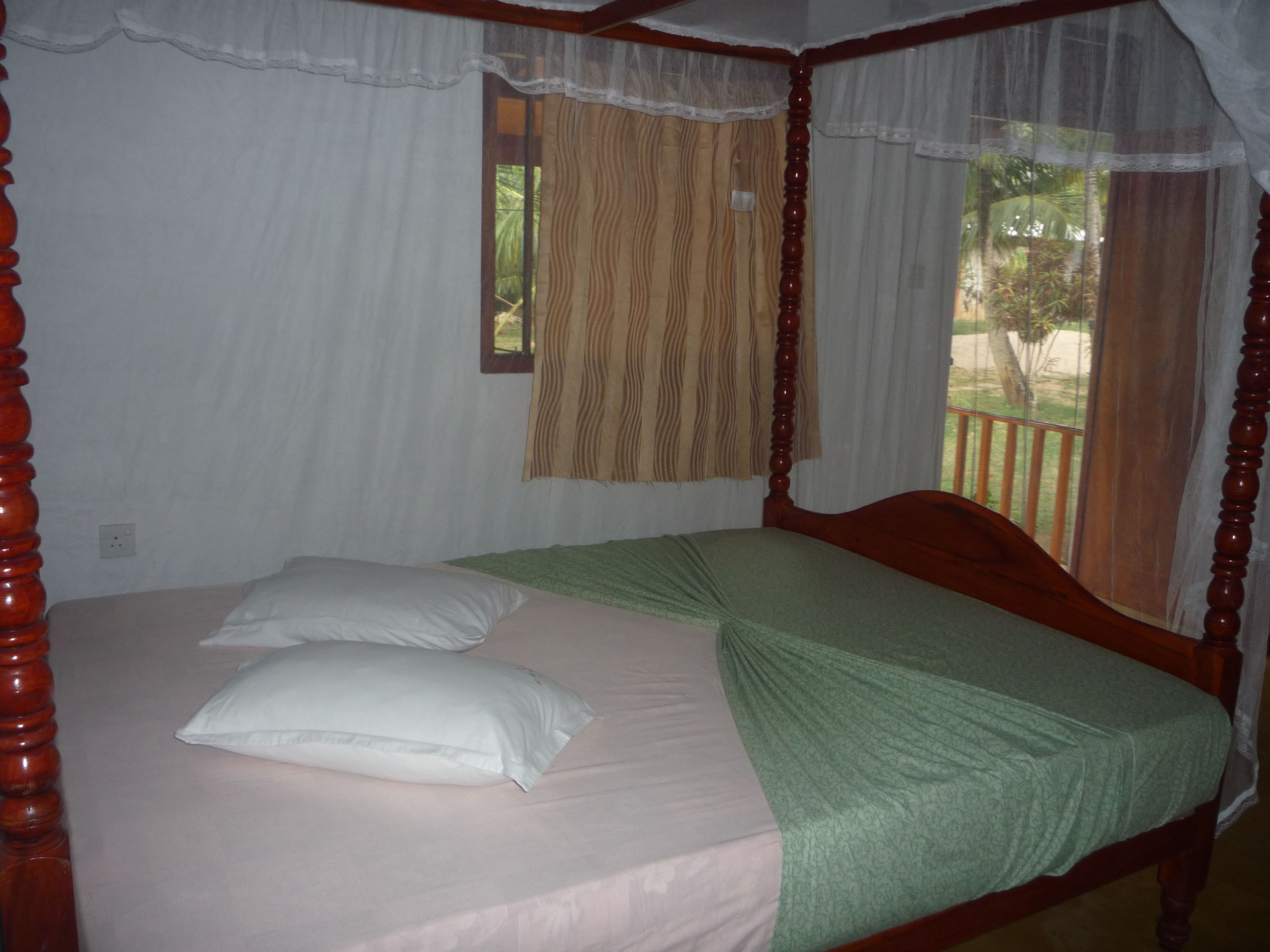 A four-poster bed with a green bedspread and white pillows, a window with a patterned curtain, and a view of greenery outside.