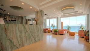 Eine Hotelhalle mit einer Marmor-Rezeption, einer Sitzecke mit Sofas, großen Fenstern mit Meerblick und einem modernen Deckenstil.