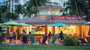 Een openluchtcafé met rieten dak, genaamd '5 o'clock Somewhere Bar', met terras en uitzicht op palmbomen.