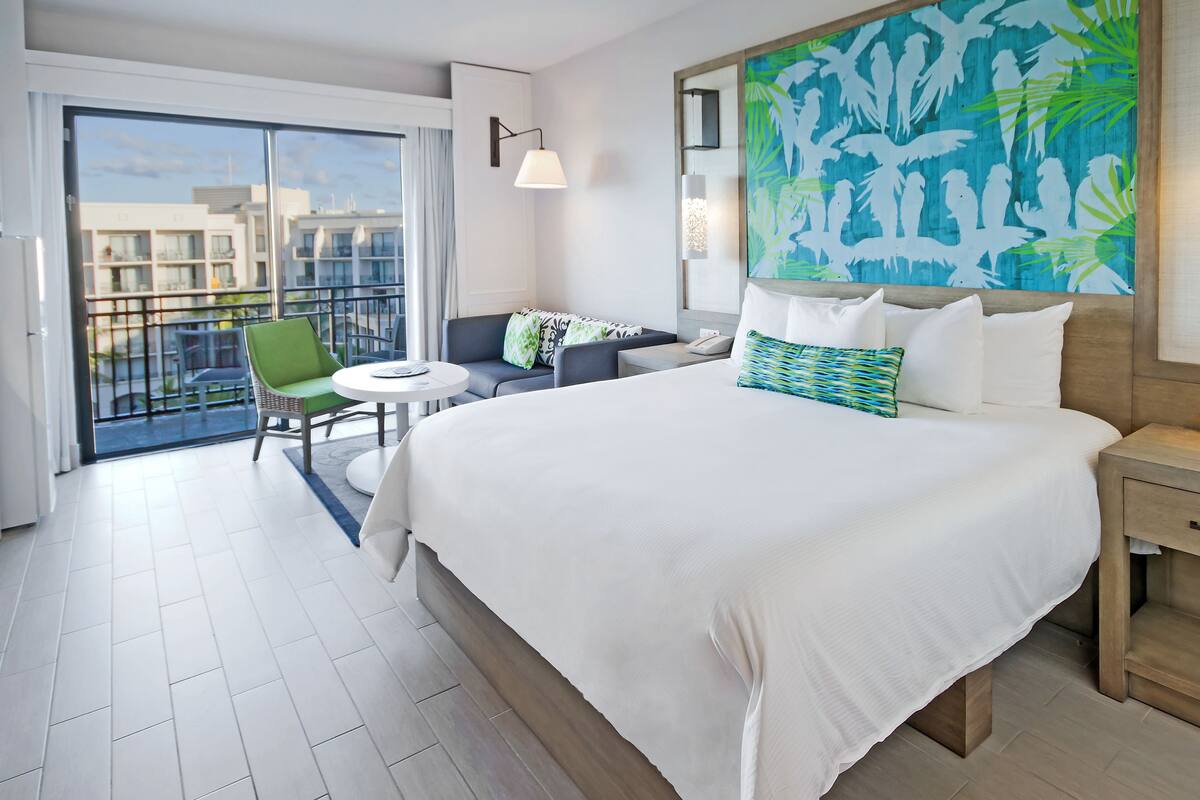 Ein Hotelzimmer mit einem großen Bett, einem kleinen runden Tisch, einem grünen Sessel, einem Balkon mit Blick auf Gebäude und einem Wandbild im tropischen Stil.