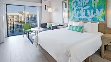 Hotelkamer met een groot bed, een klein rond tafeltje, een groene stoel, een balkon met uitzicht op gebouwen en een muurschildering met tropisch thema.