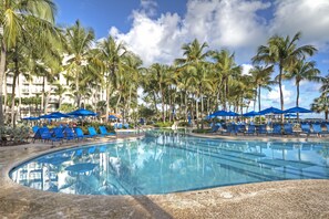 Ein Pool, umgeben von Palmen und blauen Sonnenschirmen, mit einem Gebäude im Hintergrund.