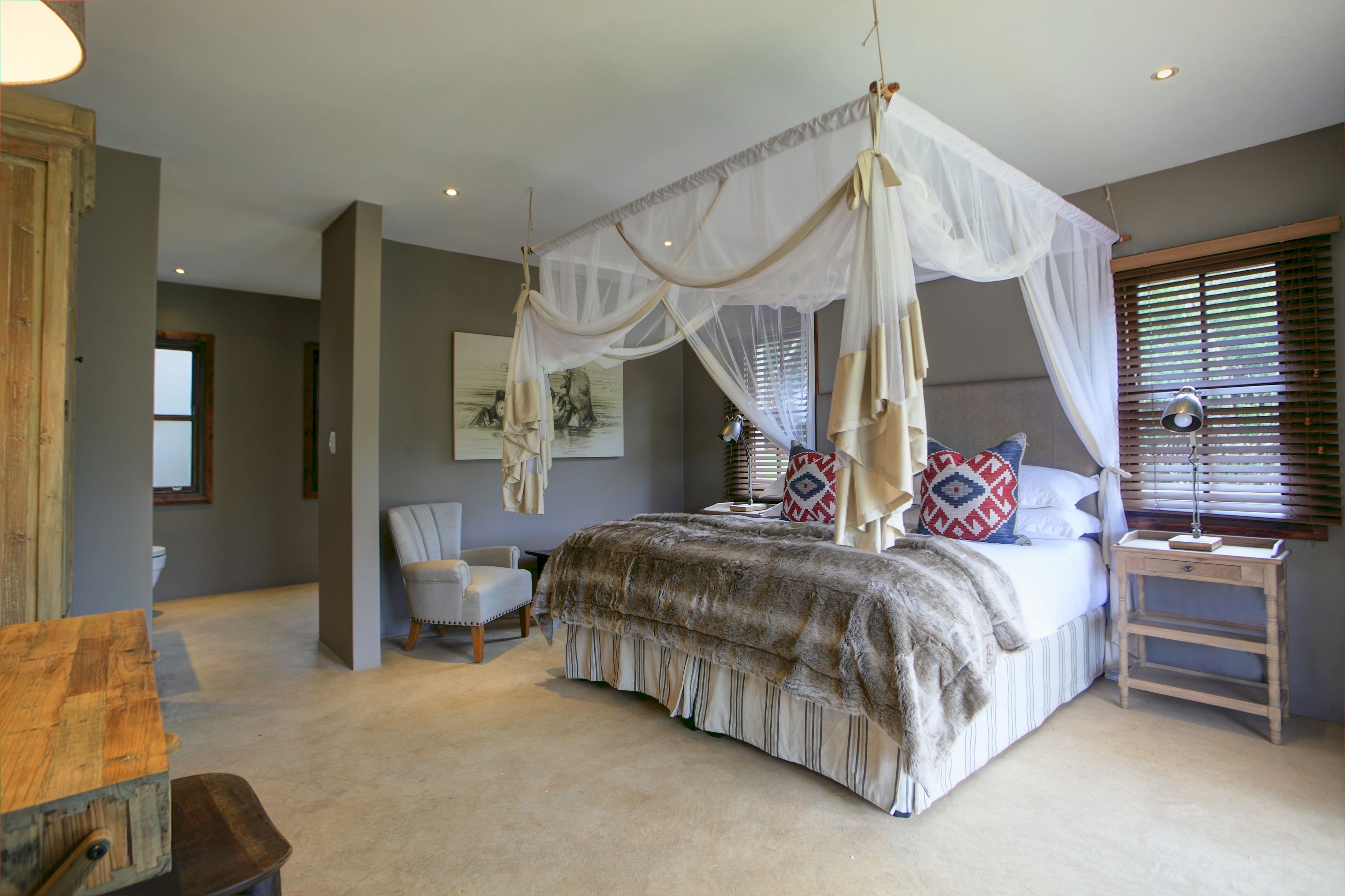 A bedroom with a four-poster bed, a chair, a bedside table, and a window with blinds.
