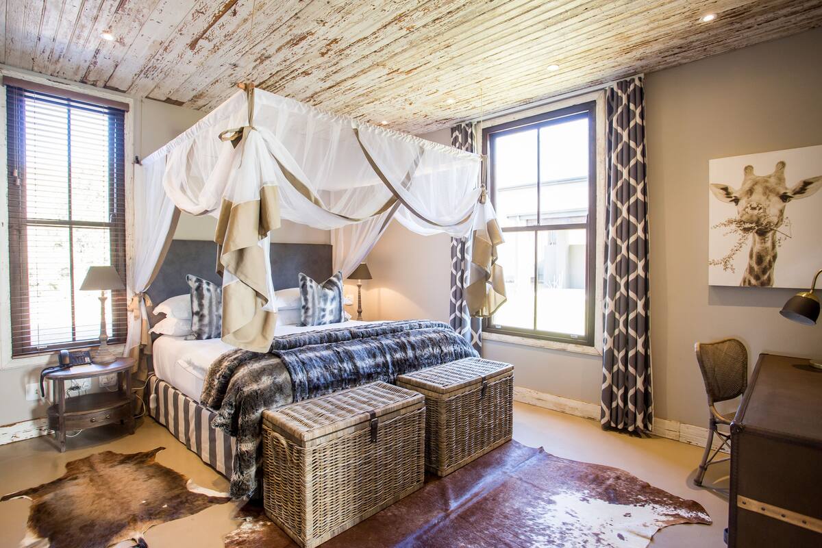 Een slaapkamer met een hemelbed, een koeienhuidtapijt, rieten meubels en een girafschilderij.