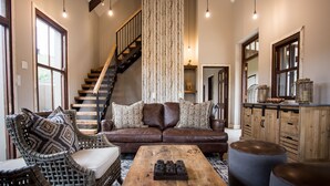 A spacious living room with a brown leather sofa, wicker chairs, a wooden coffee table, and a staircase with a wooden railing.
