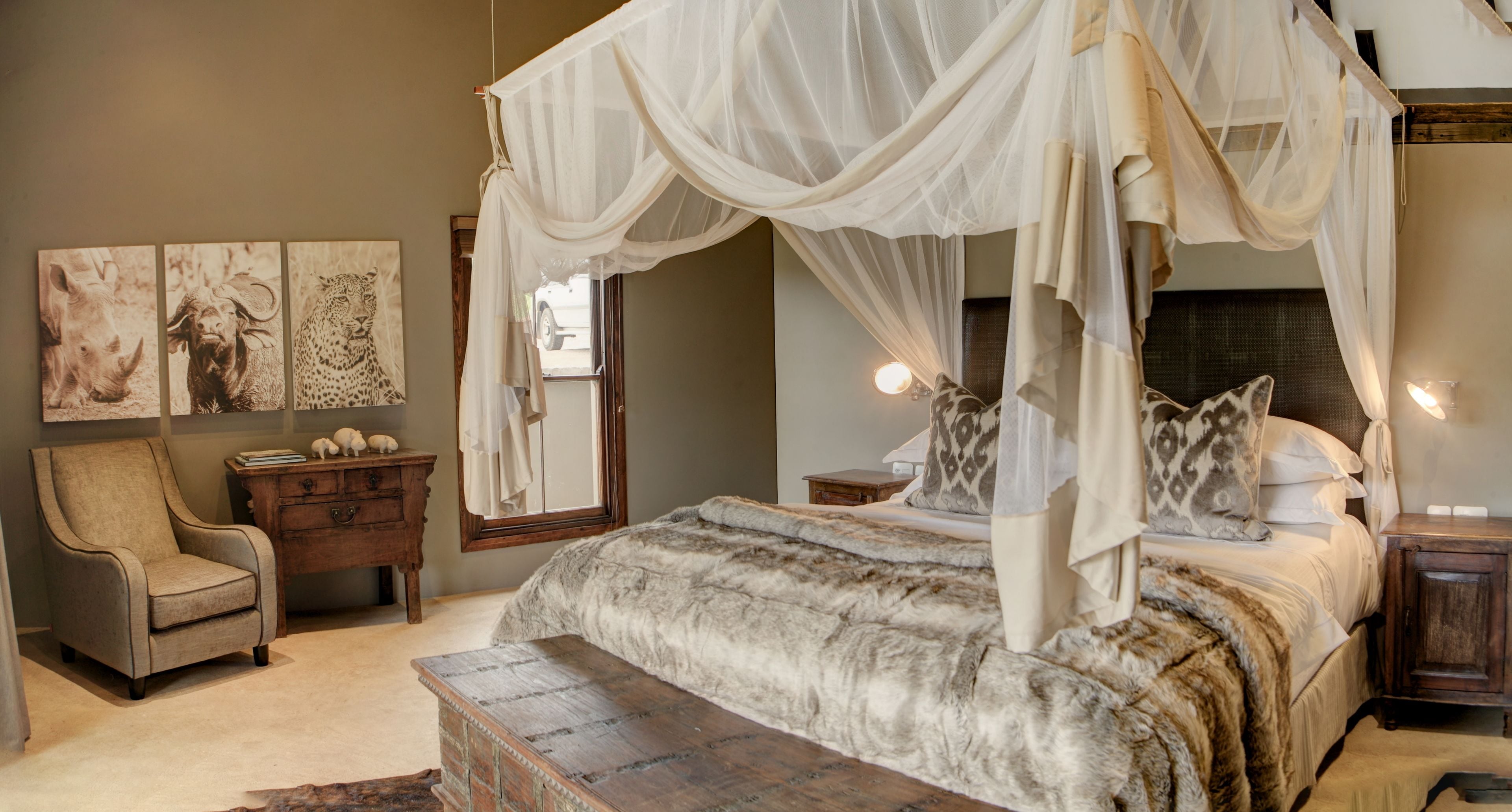 A four-poster canopy bed with a fur-like bedspread, a wooden chest, a chair, and artwork on the wall.