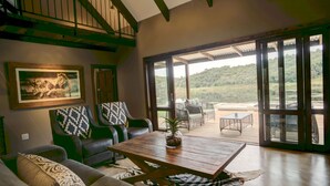 A spacious living area with a large wooden table, green upholstered chairs, and a view of a lake through large sliding glass doors.