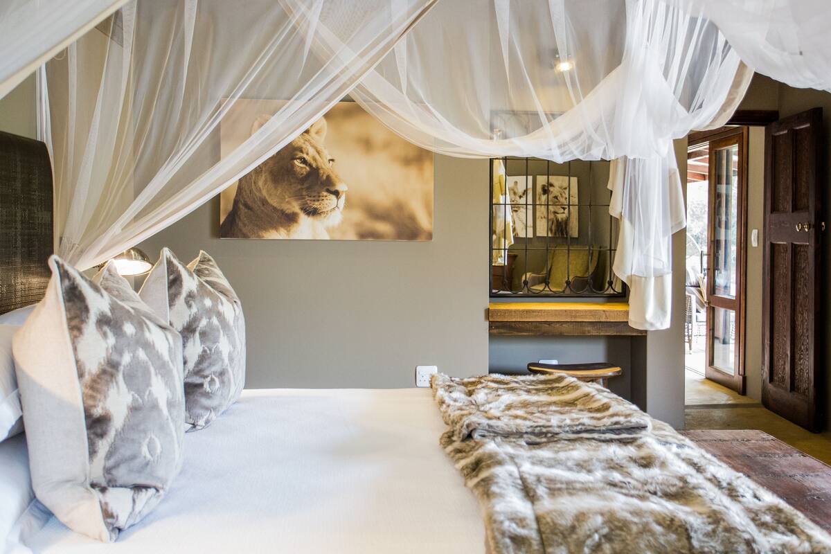 Een slaapkamer met hemelbed, schilderij van een leeuw aan de muur, een houten bankje en uitzicht naar buiten.