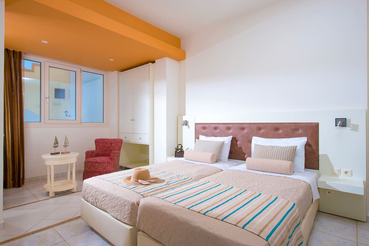 Habitación de hotel con dos camas, un sillón rojo, una mesita y un ventanal con cortinas.