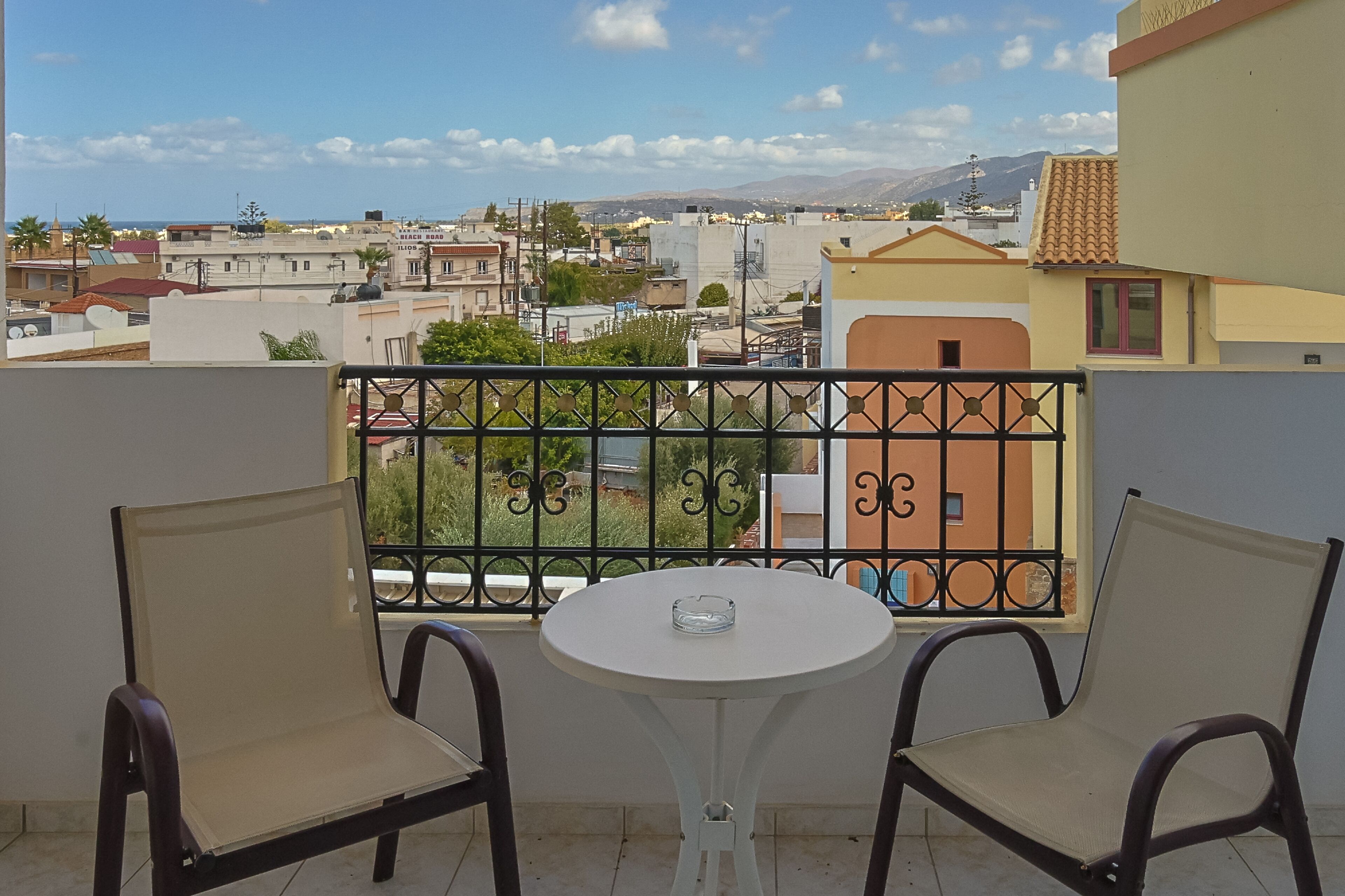 A balcony with two chairs and a small table, overlooking a residential area with buildings and mountains in the distance.