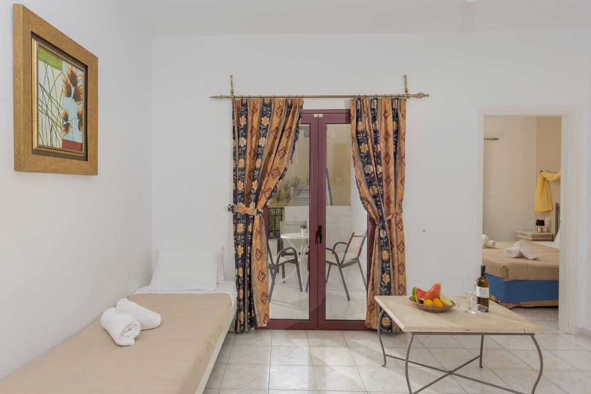 Una habitación con una cama, una mesa con fruta y una botella, y un comedor visible a través de una puerta de vidrio.