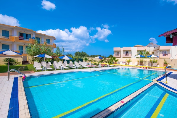 A swimming pool with lounge chairs and umbrellas, surrounded by buildings with balconies and a clear blue sky.