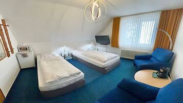 A hotel room with two single beds, a blue sofa, a small TV, a desk with a lamp, and a window with curtains.