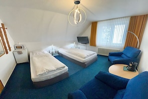 A hotel room with two single beds, a blue sofa, a small TV, a desk with a lamp, and a window with curtains.