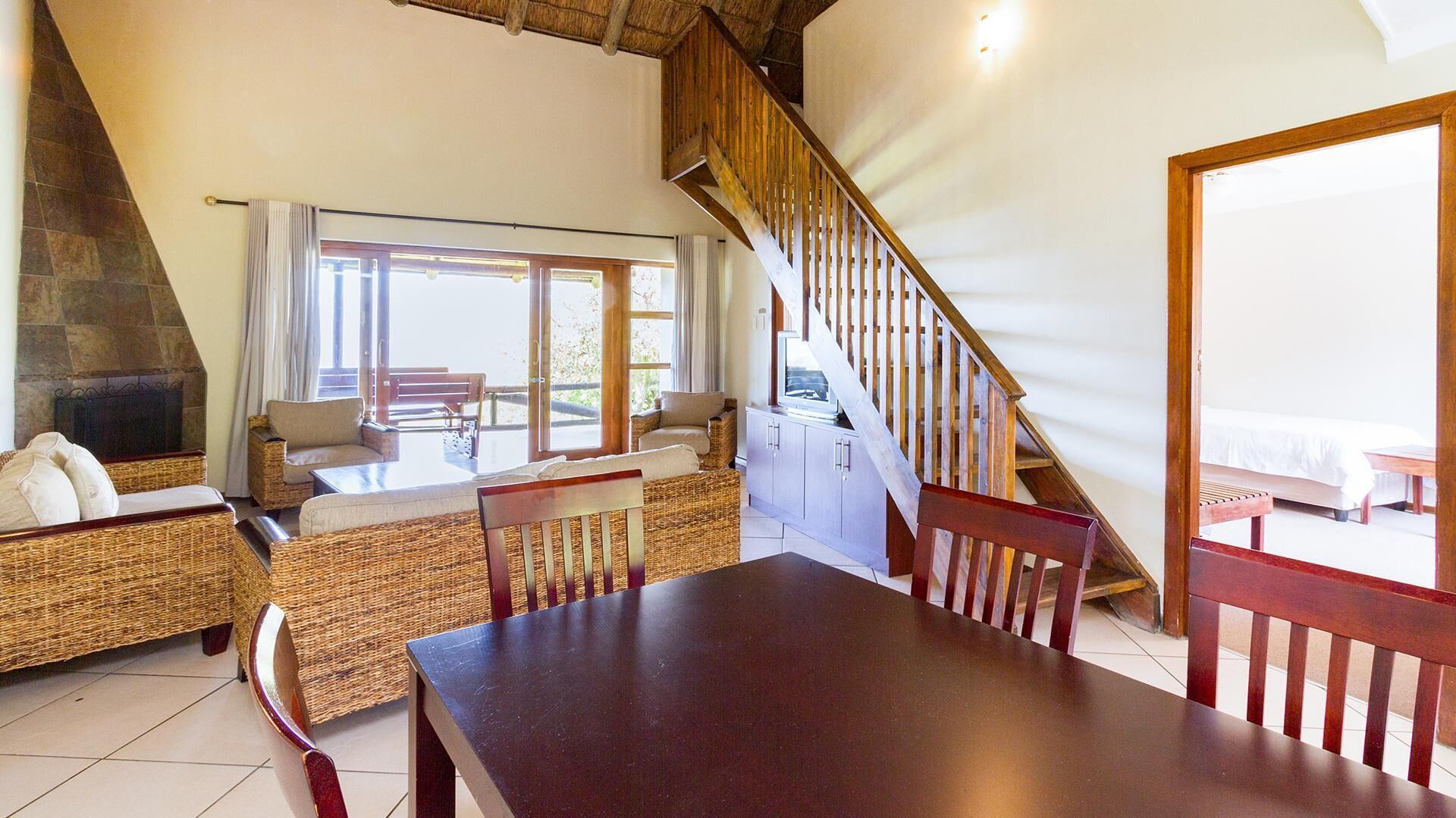 Un espace repas avec une table et des chaises en bois, une cheminée en pierre, des meubles en osier et un escalier menant à un balcon.