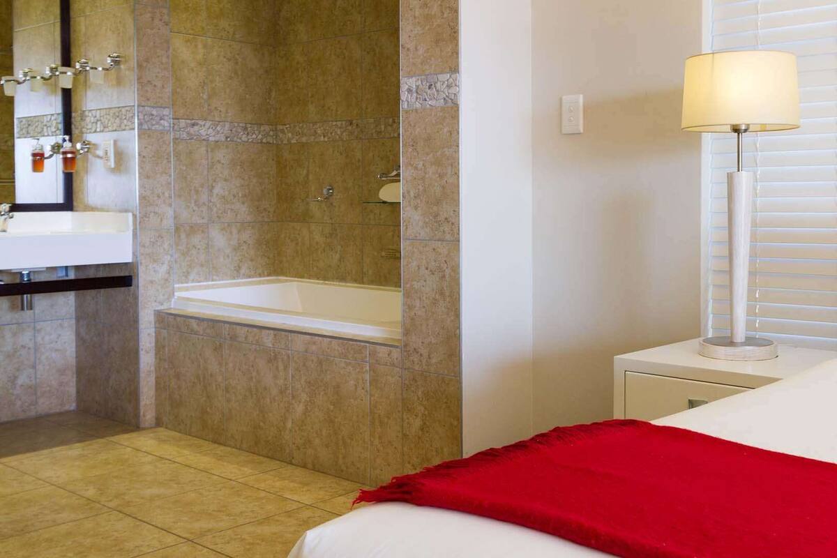 A hotel room with a bed, a nightstand, a lamp, a window with blinds, a bathroom with a sink and bathtub.