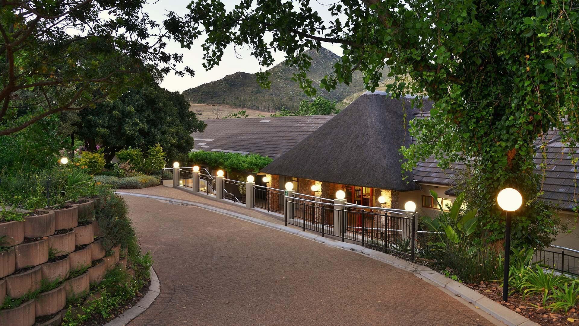 A pathway leading to a building with a thatched roof, surrounded by greenery and a mountainous backdrop.
