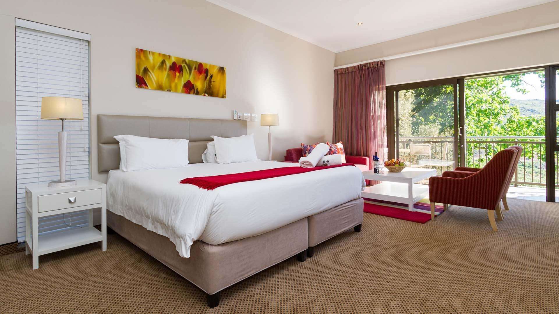 A hotel room with a large bed, two bedside tables, a desk with a chair, a window with a view of greenery, and a painting on the wall.