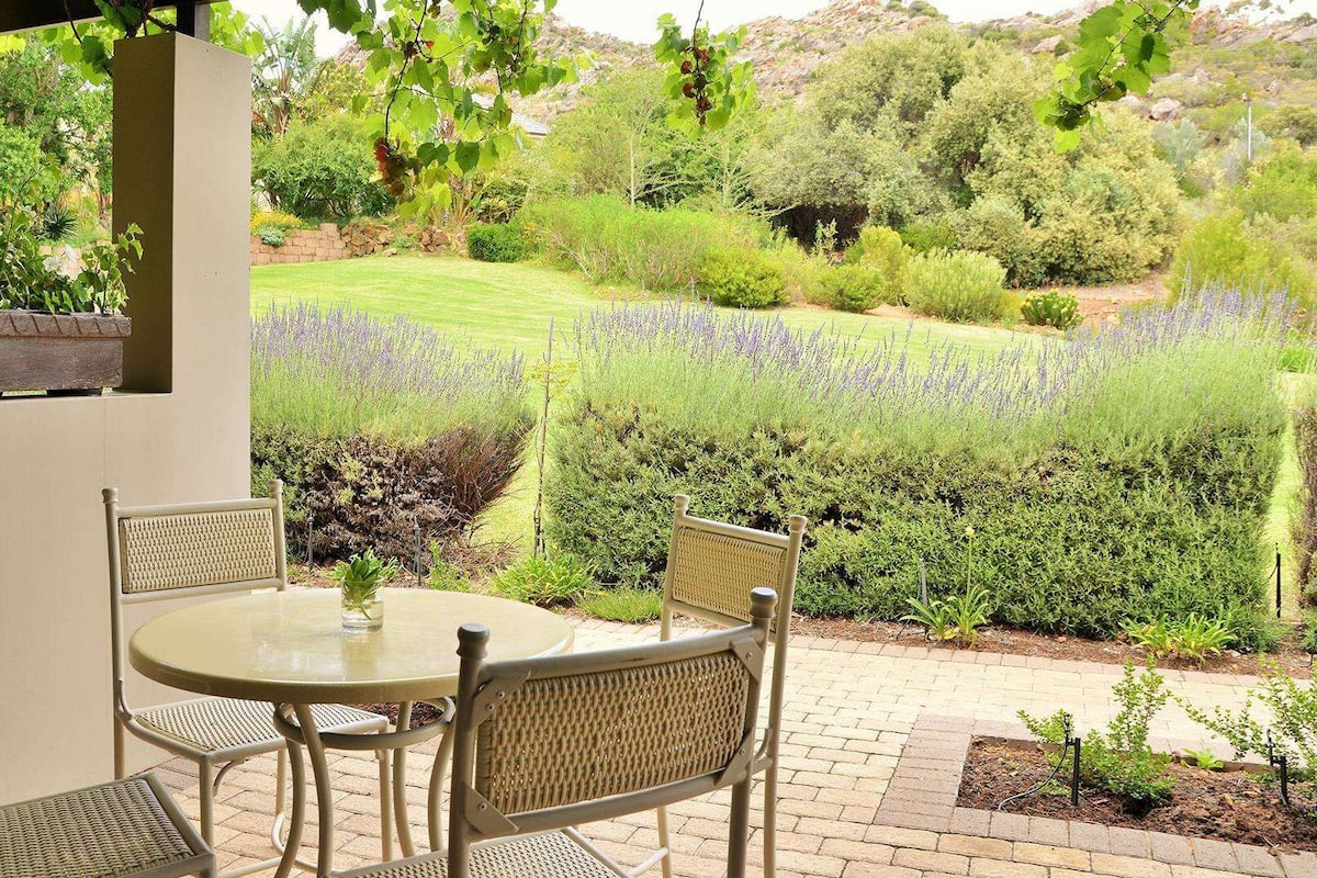 An outdoor patio with a round table and wicker chairs, surrounded by a garden with various plants and a view of a field.