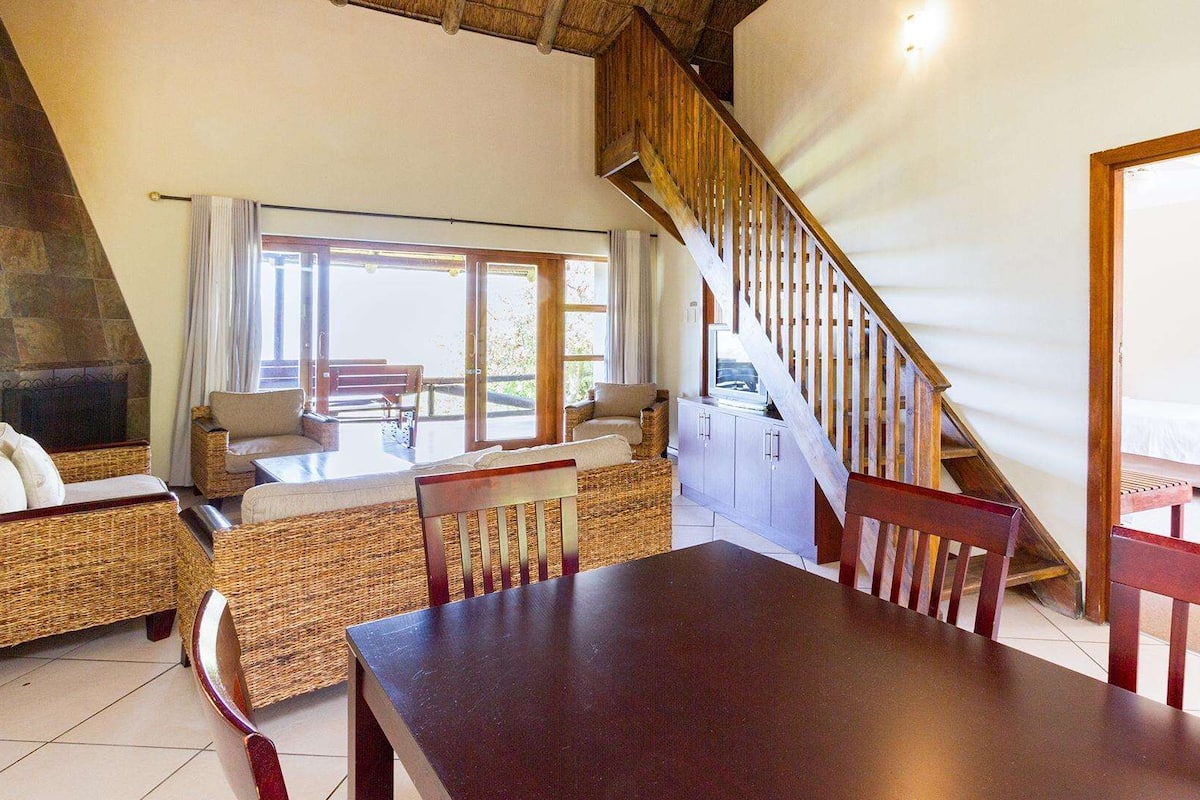 A spacious living area with a high ceiling, a wooden staircase, a fireplace, and a large window providing natural light.