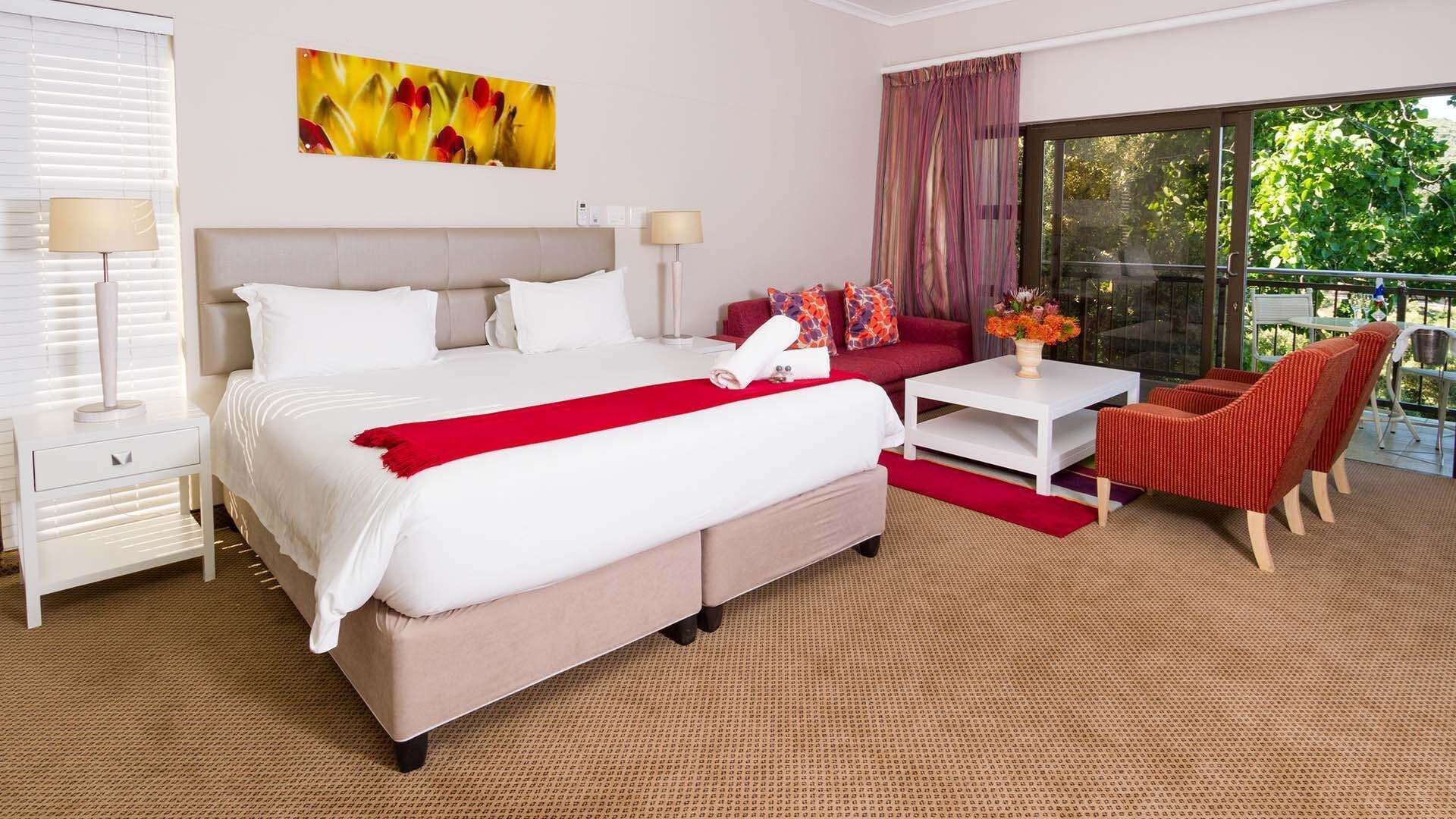 Quarto de hotel com uma cama grande, uma mesa pequena, uma cadeira vermelha e vista para o verde lá fora.