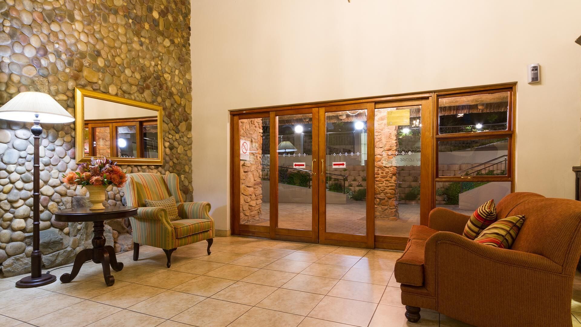 A room with a stone wall, a wooden-framed mirror, a table with a lamp, striped armchairs, and a sliding glass door leading outside.