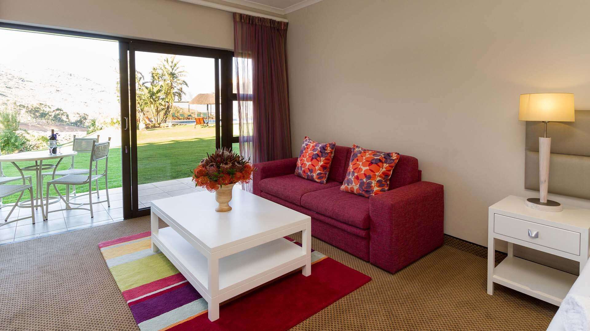 Un salon moderne avec un canapé rouge, une table basse blanche et une petite table de chevet avec une lampe. Un tapis coloré et un vase de fleurs sont posés sur la table basse.