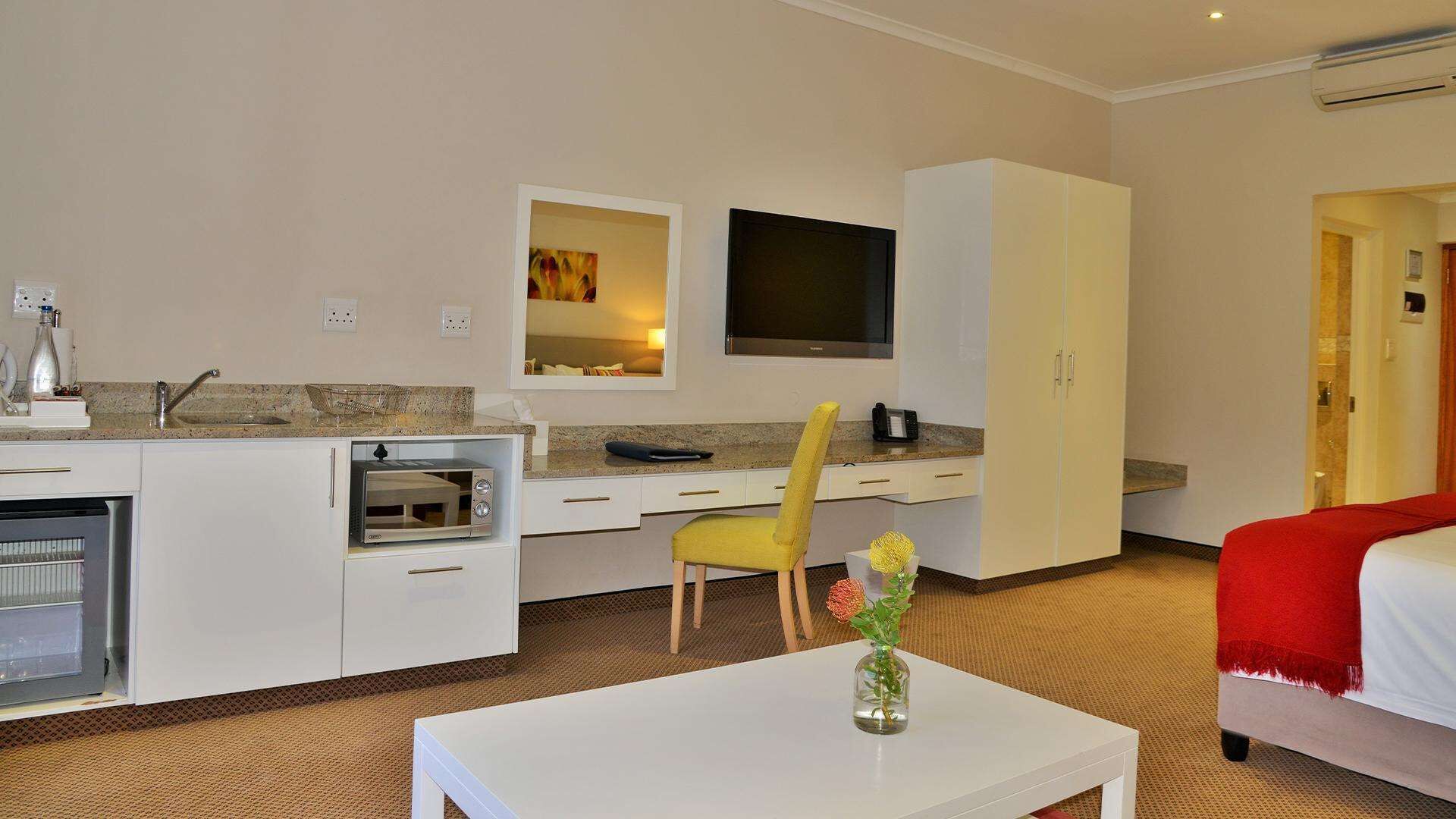 A hotel room with a kitchenette, a bed, a TV, and a small table with a vase.
