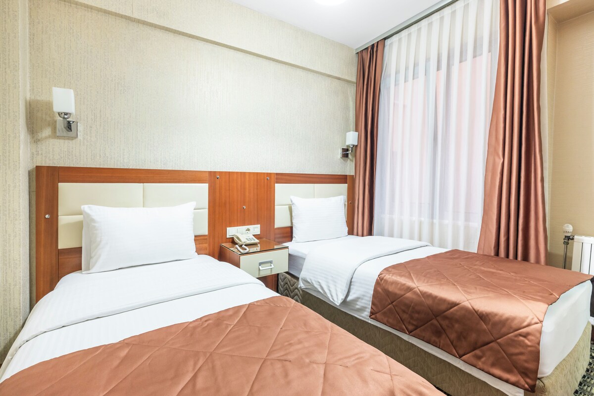 A hotel room with two beds, a wooden headboard, a bedside table with a phone, and a window with curtains.