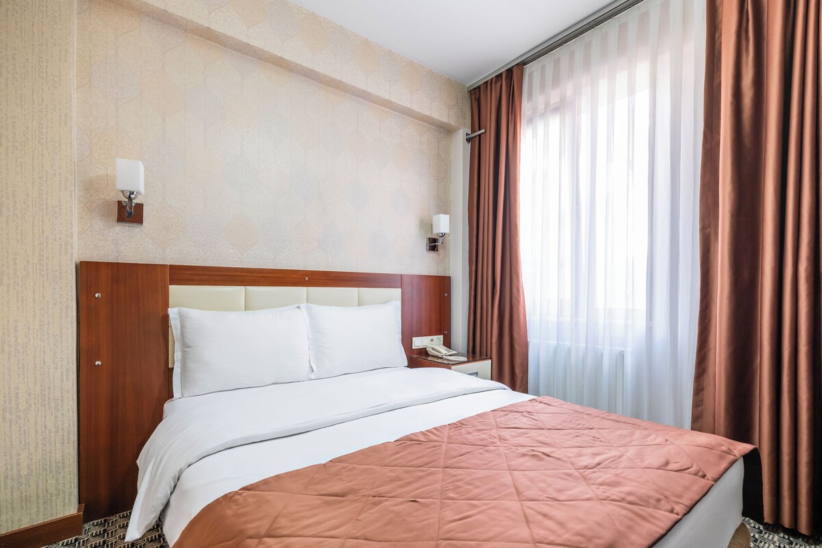 A hotel room with a large bed, a wooden headboard, a bedside table with a phone, and a window with curtains.