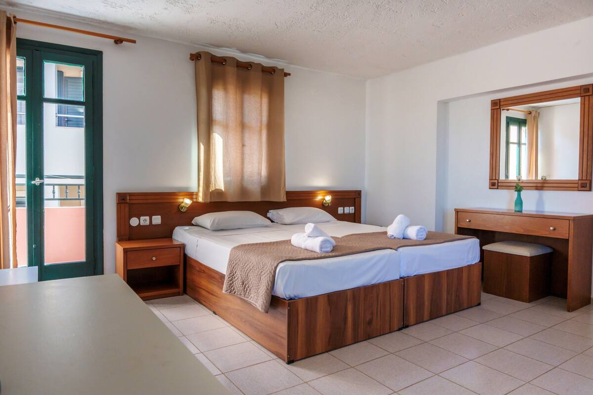 A hotel room with a wooden bed, a bedside table, a nightstand, a desk with a mirror, and a view of the outside through a door.