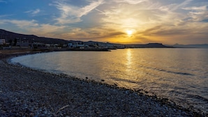 Escena costera con una playa de guijarros, un mar tranquilo y un atardecer en el horizonte.