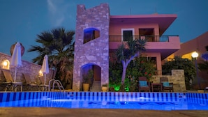 Un área de piscina con una estructura de piedra, sillones de descanso y un edificio de dos pisos con balcones.