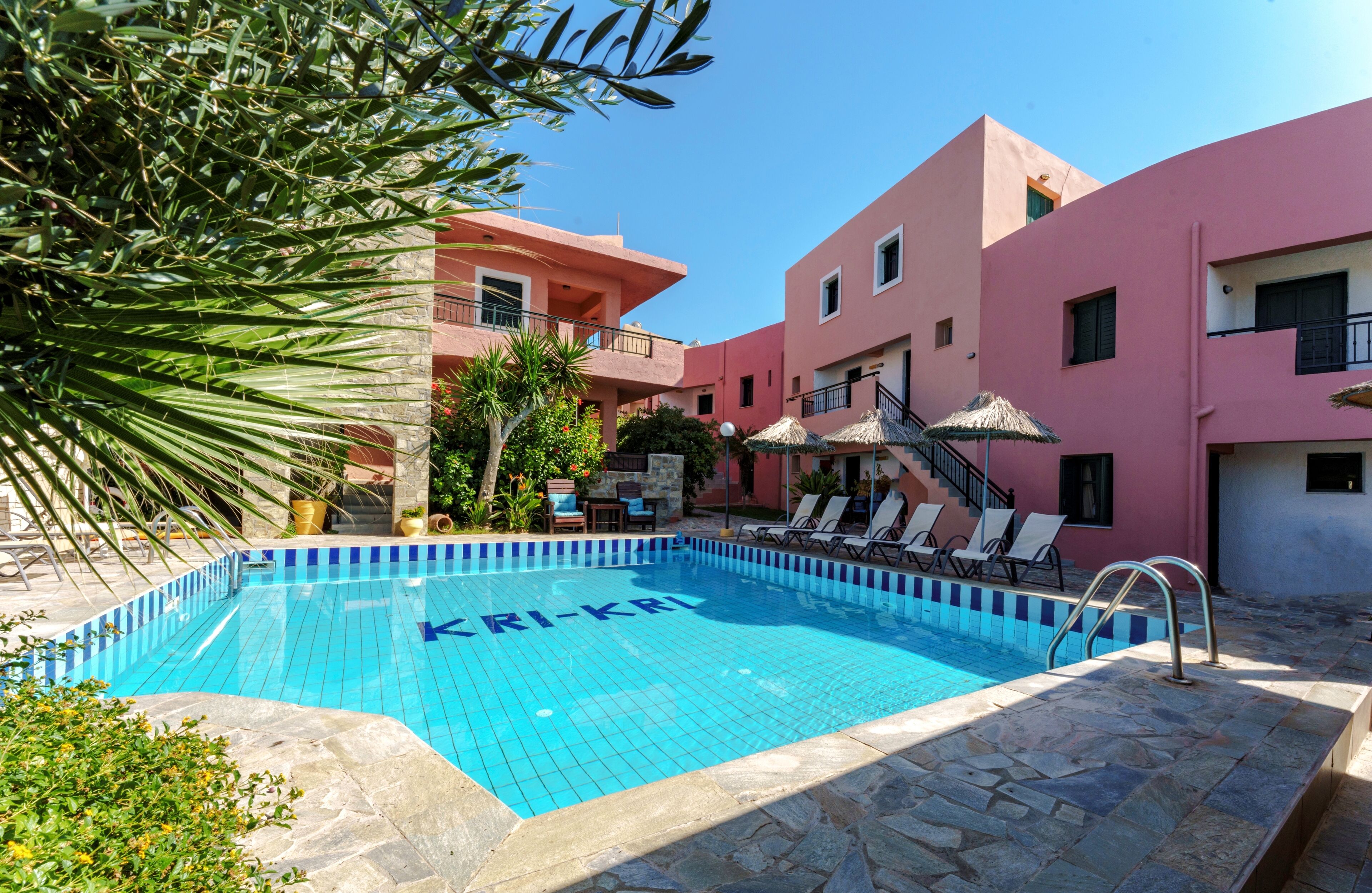A swimming pool with 'KRI-KRI' written on the water, surrounded by stone tiles, with lounge chairs and umbrellas.