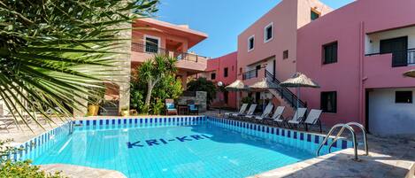 A swimming pool with 'KRI-KRI' written on the water, surrounded by stone tiles, with lounge chairs and umbrellas.