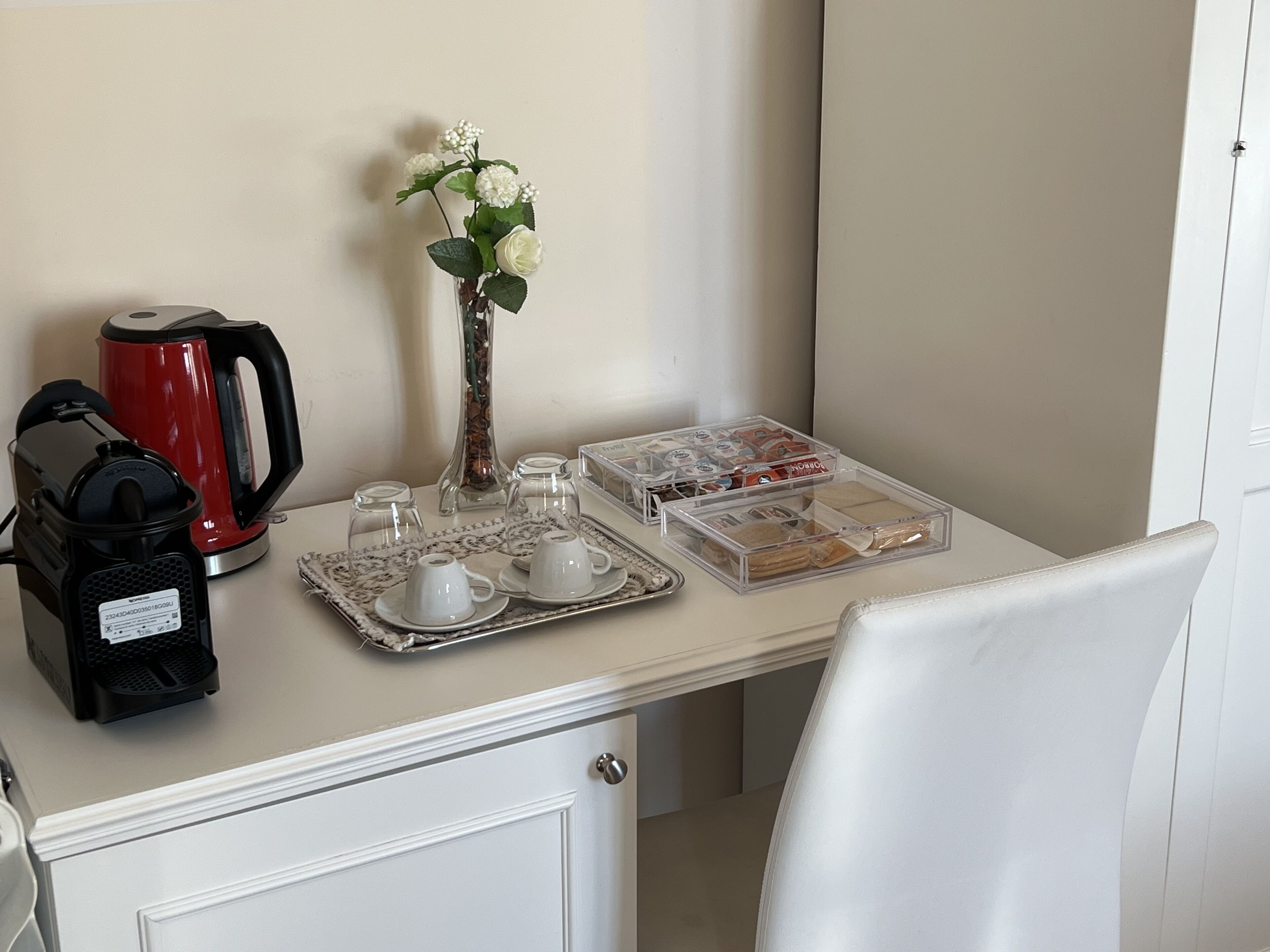 Una presentación ordenada de desayuno con cafetera, tetera, tazas y una bandeja de pasteles sobre una mesa blanca.