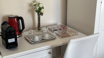 Una presentación ordenada de desayuno con cafetera, tetera, tazas y una bandeja de pasteles sobre una mesa blanca.