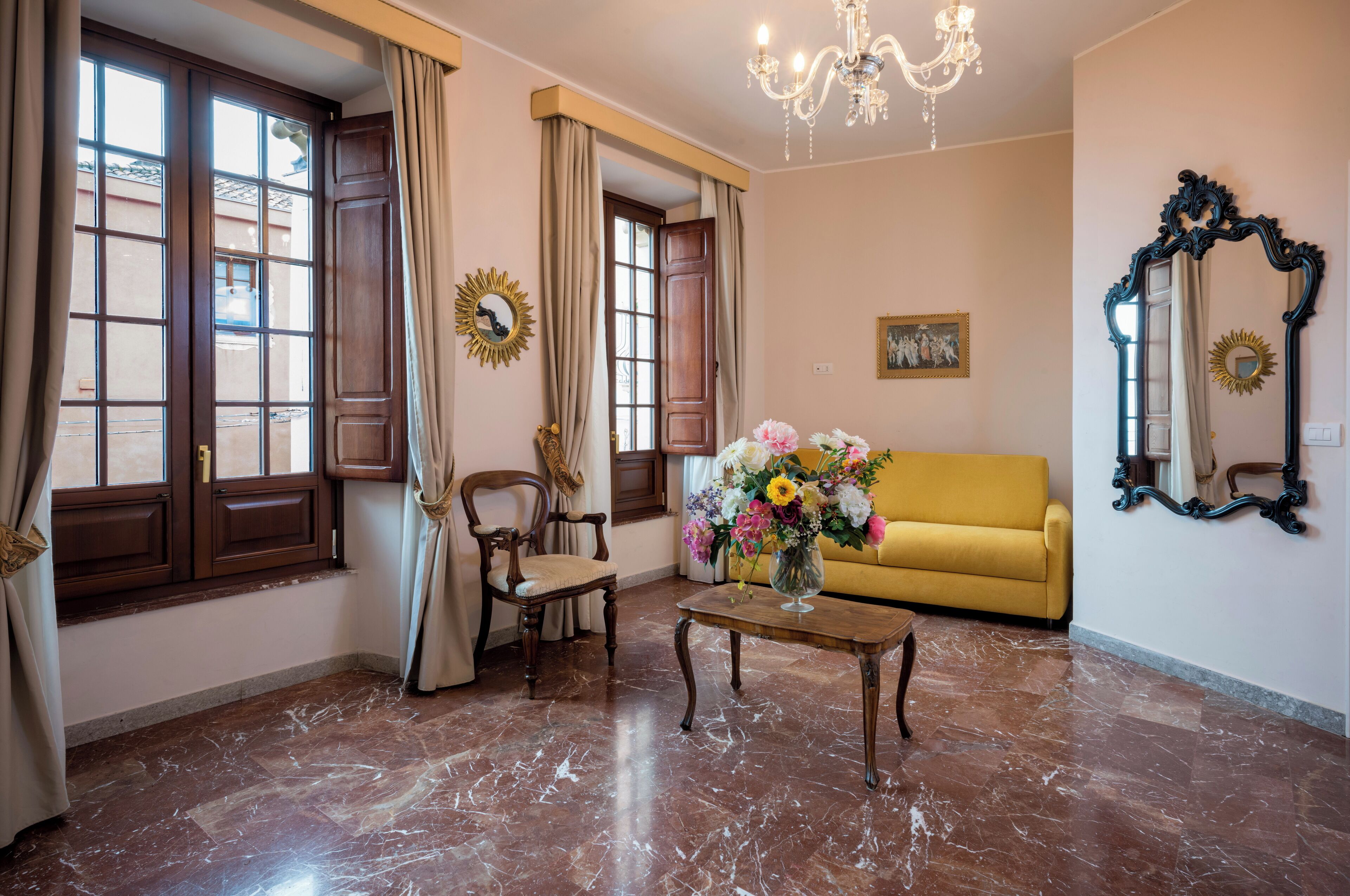 A living room with a yellow sofa, a wooden chair, a coffee table with a vase of flowers, a chandelier, and a large mirror on the wall.