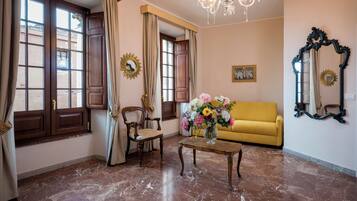 A living room with a yellow sofa, a wooden chair, a coffee table with a vase of flowers, a chandelier, and a large mirror on the wall.