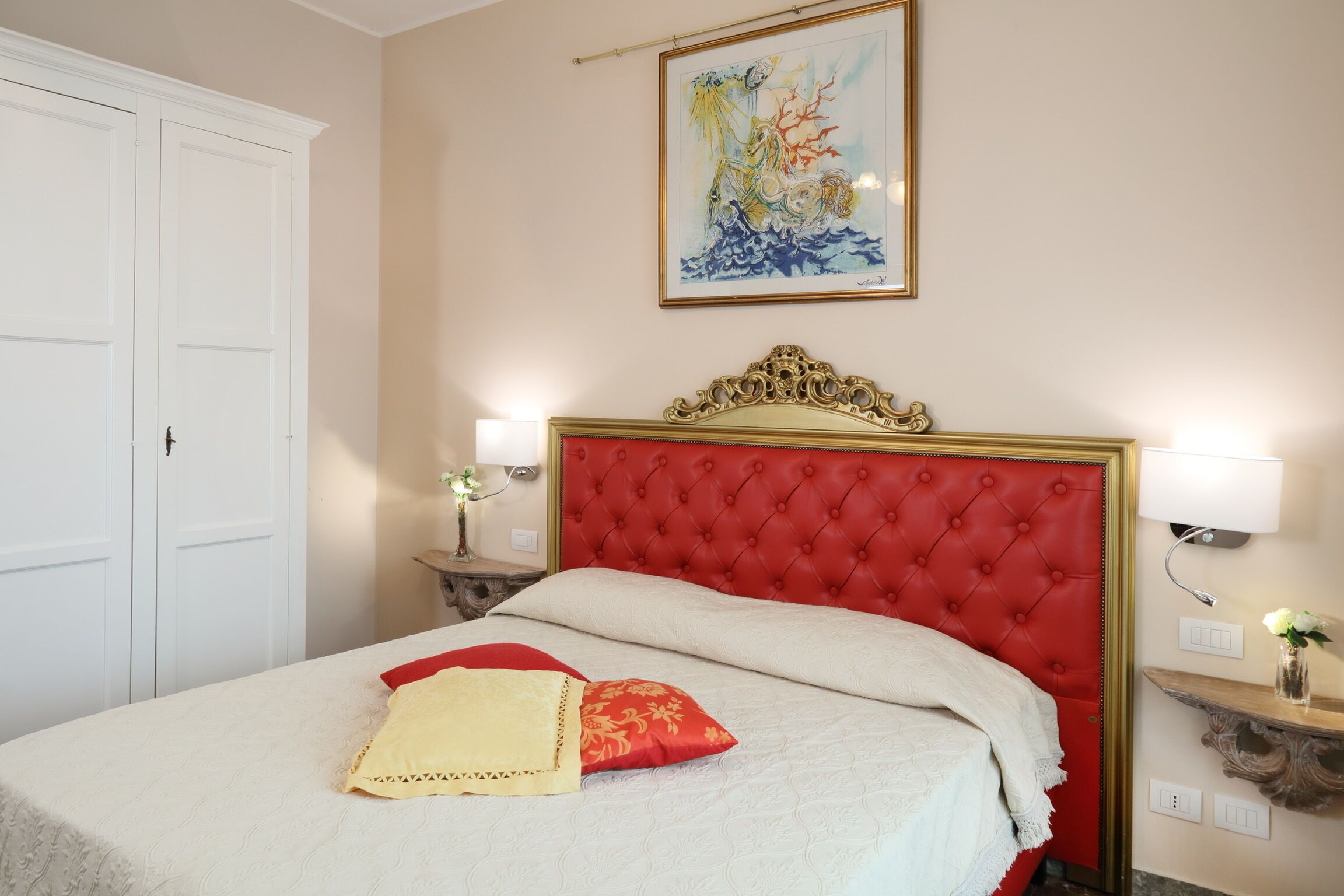 A bedroom with a red upholstered headboard, a white bedspread, and a single red and yellow pillow. There is a painting on the wall above the bed and a small table with a vase of flowers to the right.