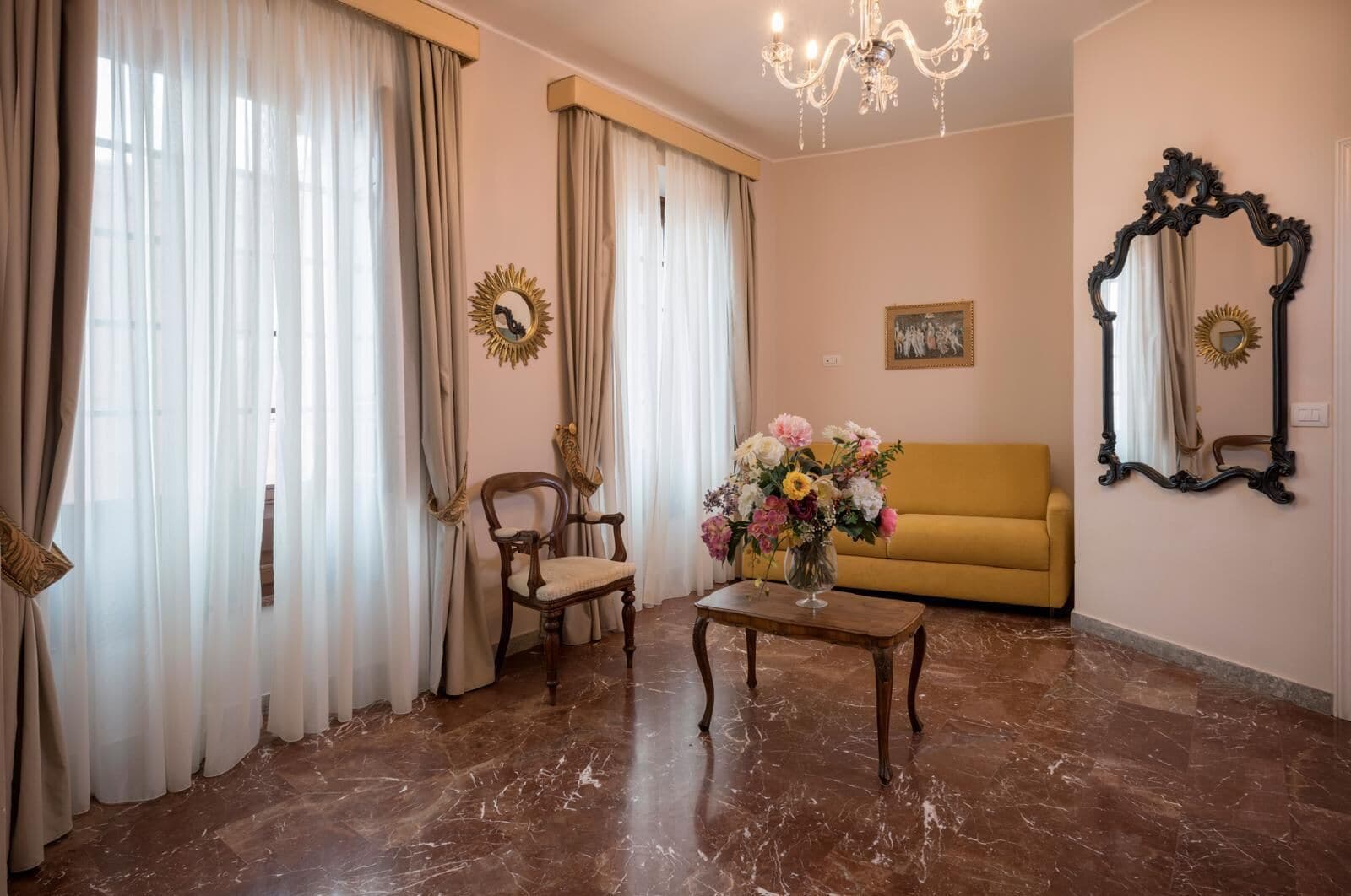 Ein Wohnzimmer mit einer Couch, einem Sessel, einem Tisch mit Blumenvase, einer Chandelüster und zwei Wandspiegeln.